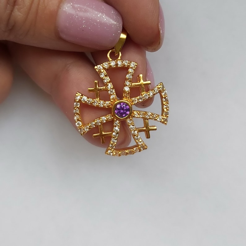 May include: A gold pendant with a purple gemstone in the center. The pendant is in the shape of a cross with smaller crosses around the edges. The pendant is surrounded by small clear stones.