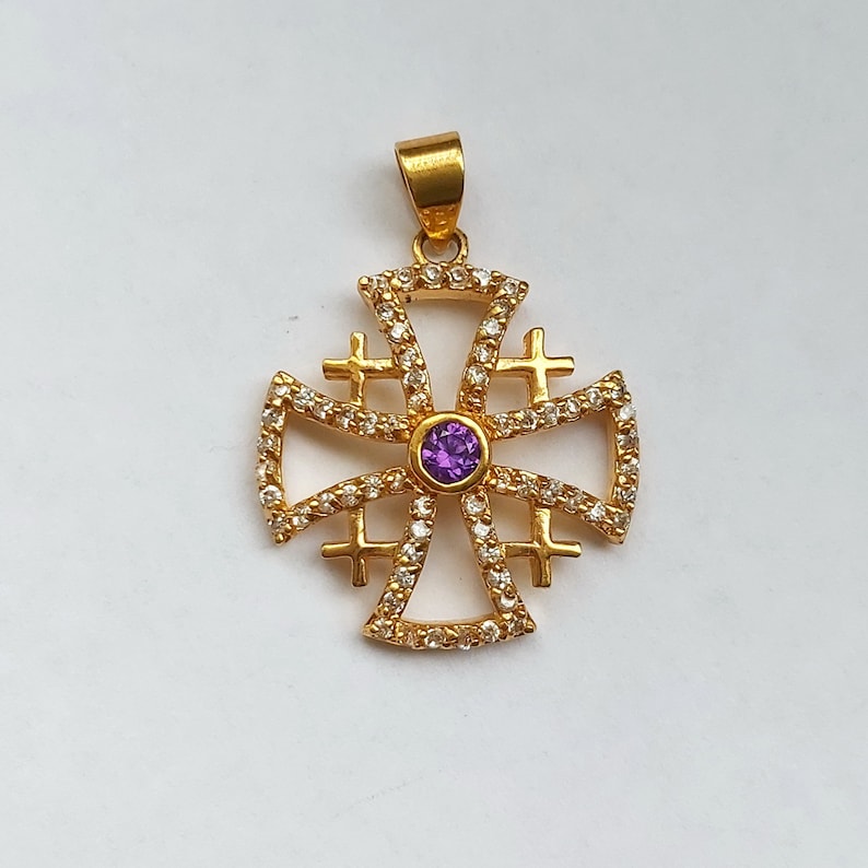 May include: A gold pendant in the shape of a cross with a purple gemstone in the center. The cross is decorated with smaller white gemstones.