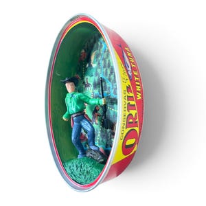 Könnte beinhalten: Eine umfunktionierte ovale Metalldose von Ortiz White Tuna mit einer Miniatur-Cowboyfigur in grünem Hemd und blauer Jeans, die ein Gewehr hält. Das Innere der Dose zeigt eine aufgedruckte Waldszene mit einem kleinen Tier und einem Reiter.