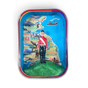 Puede incluir: Una lata de metal rectangular que contiene una figura en miniatura de un soldado con túnica roja y falda escocesa verde sobre un montículo verde. El fondo presenta un mapa colorido de Escocia con nombres de lugares como Edimburgo y Perth.