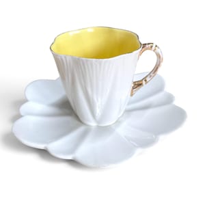 Op de afbeelding: Een witte porseleinen kop en schotel set met een geschulpt, bloembladachtig ontwerp. De binnenkant van de kop is zacht pastelgeel en het oortje heeft een goudkleurige afwerking met een decoratief, gestructureerd patroon langs de rand.