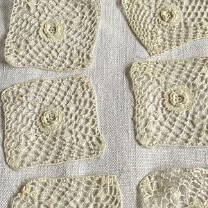 Op de afbeelding: Een verzameling vierkante, crèmekleurige gehaakte onderzetters met een bloemmotief in het midden. De onderzetters liggen op een lichte linnen achtergrond. Het open, kantachtige haakwerk geeft een vintage uitstraling, ideaal voor woondecoratie.