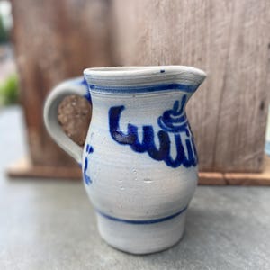 Puede incluir: Jarra pequeña de cerámica blanquecina con asa curva. La jarra presenta letras pintadas en azul y detalles decorativos. Una banda azul rodea la parte superior e inferior. La jarra es probablemente hecha a mano y tiene una estética rústica.