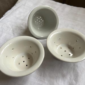 Può includere: Tre colini da tè in ceramica bianca con una finitura liscia e lucida. Ogni colino a forma di ciotola presenta un bordo largo e una serie di piccoli fori di drenaggio distanziati uniformemente sul fondo e lungo le pareti interne.