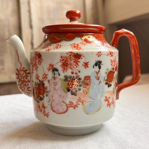 May include: A white porcelain teapot featuring a vibrant orange lid and handle. The body is decorated with hand-painted figures in traditional attire, surrounded by intricate orange floral patterns and gold accents on the spout and rim.