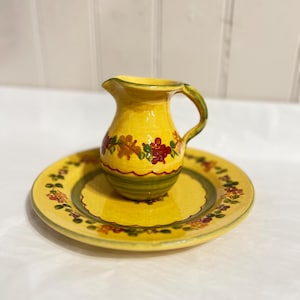 Puede incluir: Una jarra pequeña de cerámica amarilla con un plato a juego. La jarra y el plato están decorados con motivos florales en rojo, verde y amarillo. La jarra tiene un asa y un pico. El plato es redondo.
