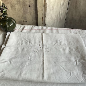 May include: A folded white textile sheet featuring intricate white embroidered monogram initials in the center. The fabric has a smooth, matte appearance with visible folds and creases, showcasing a clean, neutral aesthetic for home linens.