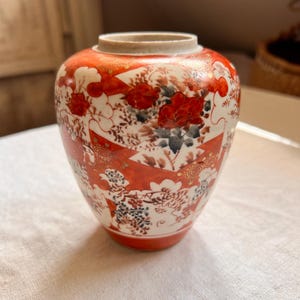 Könnte beinhalten: Eine kleine Keramikvase mit einer leuchtend orange-weißen Farbpalette, verziert mit komplexen Blumenmustern und goldfarbenen Akzenten. Das Gefäß hat einen abgerundeten Körper, einen kurzen Hals und eine weite Öffnung mit botanischen Motiven.