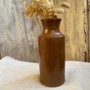 May include: A brown ceramic vase with a narrow neck and a wide base, filled with dried wheat stalks. The vase has a smooth, glossy finish and stands on a white cloth. The background is a weathered, light-colored wall.