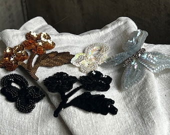 May include: Four decorative fabric appliques featuring floral and butterfly designs. The appliques are made of sequins and beads in shades of gold, black, white, and iridescent blue.