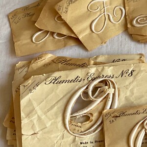 May include: A stack of vintage paper packets with white embroidered letters and the text "Plumetis-Express N:8" and "Plumetis-Express N:4". The packets are labeled with the price "25F" and "20F".