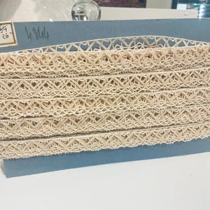 May include: A spool of beige lace trim featuring a repeating geometric diamond pattern, wrapped around a rectangular blue cardboard card. The card displays handwritten text including the numbers 4754, 26, and 4 3/4.