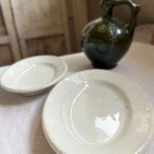Könnte beinhalten: Zwei weiße, ovale Keramikschalen mit glatten, glänzenden Oberflächen und erhöhtem Rand, begleitet von einem dunkelgrün glasierten Keramikkrug mit Henkel, engem Hals und einem dekorativen Muster aus kleinen weißen Punkten am bauchigen Körper.