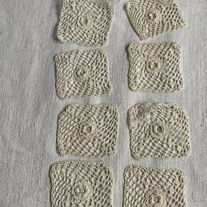 Op de afbeelding: Acht vierkante, crèmekleurige gehaakte onderzetters met een bloemmotief in het midden. De onderzetters zijn gerangschikt op een getextureerd, lichtgekleurd oppervlak, wat hun delicate, handgemaakte uiterlijk benadrukt. Ideaal voor vintage decoratie.