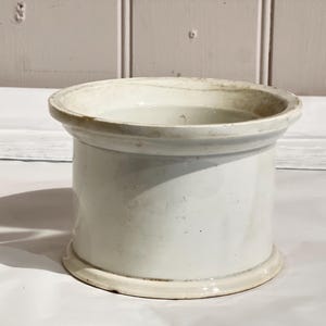 May include: A white ceramic jar with a cylindrical body and a wide, slightly flared rim. The jar has a simple, classic design and appears to be vintage. The surface has subtle variations in colour, suggesting age and use. The jar is suitable for kitchen or bathroom storage.