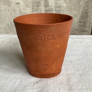 May include: A terracotta-colored flower pot with a slightly flared shape. The pot has a textured surface and a visible inscription around the top rim. The pot is sitting on a white surface.