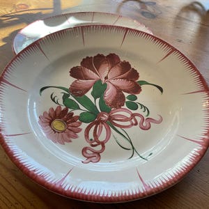 May include: Two white ceramic plates with a floral design. The plates have a red rim with a scalloped edge. The floral design features a large maroon flower, a smaller pink flower, green leaves, and a red ribbon.