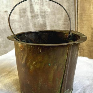 Könnte beinhalten: Ein kleiner, verwitterter Kupferkübel mit dunkler, oxidierter Patina und sichtbarem Grünspan. Der Kübel verfügt über einen dünnen, gebogenen Metalldrahtgriff, der an einem breiten, flachen Rand befestigt ist, sowie eine vertikale Naht.