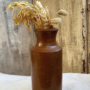 Puede incluir: Un jarrón de cerámica marrón con cuello estrecho y base ancha, lleno de espigas de trigo secas. El jarrón tiene un acabado brillante y está sobre una superficie blanca. El fondo presenta paneles de madera desgastados.