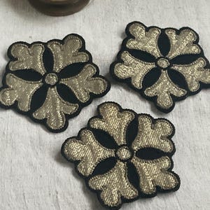 May include: Three decorative embroidered appliques. Each applique features a gold-colored, floral-like design on a black background. The appliques are arranged on a white surface.