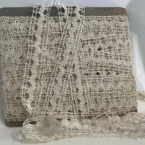 May include: A spool of off-white lace trim featuring a repeating scalloped edge and an openwork mesh pattern. The delicate fabric is wound around a rectangular cardboard base, with a loose end extending forward to show the intricate weave.