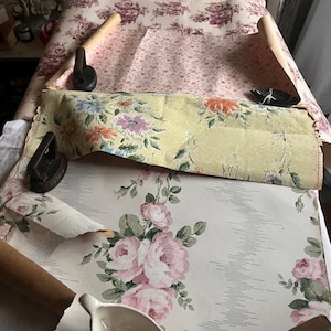 May include: Three rolls of vintage wallpaper with floral patterns. The wallpaper is in various colors, including pink, green, and yellow. The rolls are on a white surface with a small white pitcher.