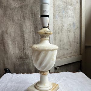 May include: A white marble table lamp with a bulb. The lamp has a square base and a decorative, twisted urn-shaped body. The lamp is off-white with some tan accents. The lamp is on a white surface.