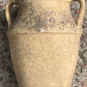May include: A beige-coloured amphora with two curved handles. The ceramic vessel has a textured surface and shows signs of age, with darker patches and wear. The amphora's shape tapers towards the base, suggesting it was used for storage.