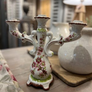 Könnte beinhalten: Ein weißer Kerzenständer aus Keramik mit drei Halterungen, verziert mit einem handgemalten Blumenmuster aus rosa und roten Blüten sowie grünen Blättern. Der Fuß und die Ränder sind mit einem dunkelroten Saum akzentuiert und die Arme geschwungen.