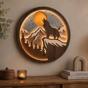 Puede incluir: Una pieza de arte mural circular de madera con una silueta en capas de un lobo aullando a una luna brillante sobre picos de montañas y pinos. El diseño está retroiluminado, proyectando una luz cálida que resalta los recortes de madera oscura.