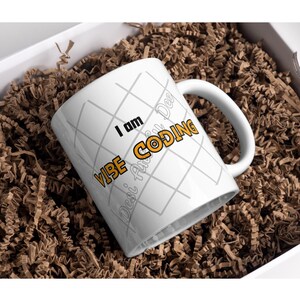 May include: White ceramic mug with the text "I am Vibe Coding" in black and orange, with a grey grid pattern. The mug is inside a white box filled with brown crinkle paper.