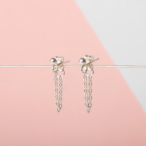May include: A pair of silver earrings with dangling chains. The earrings are on a pink and white background.