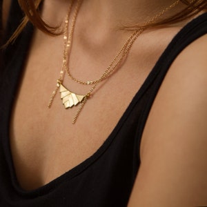 May include: Gold-coloured layered necklace with a geometric fan-shaped pendant. The necklace has two delicate chains and small dangling accents. The jewellery is displayed against a dark background, highlighting its metallic shine.