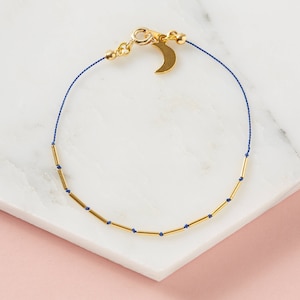 May include: A gold bracelet with a crescent moon charm and small gold beads on a blue cord.