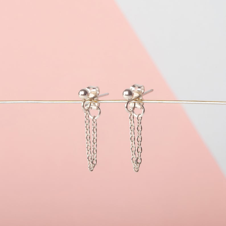 May include: A pair of silver earrings with dangling chains. The earrings are on a pink and white background.