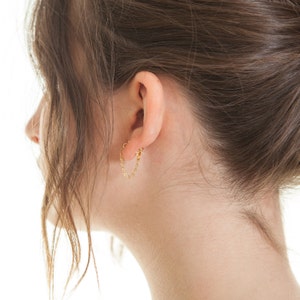 May include: A close-up of a person's ear with a gold chain earring. The chain is looped through the earlobe and then through a small hoop.