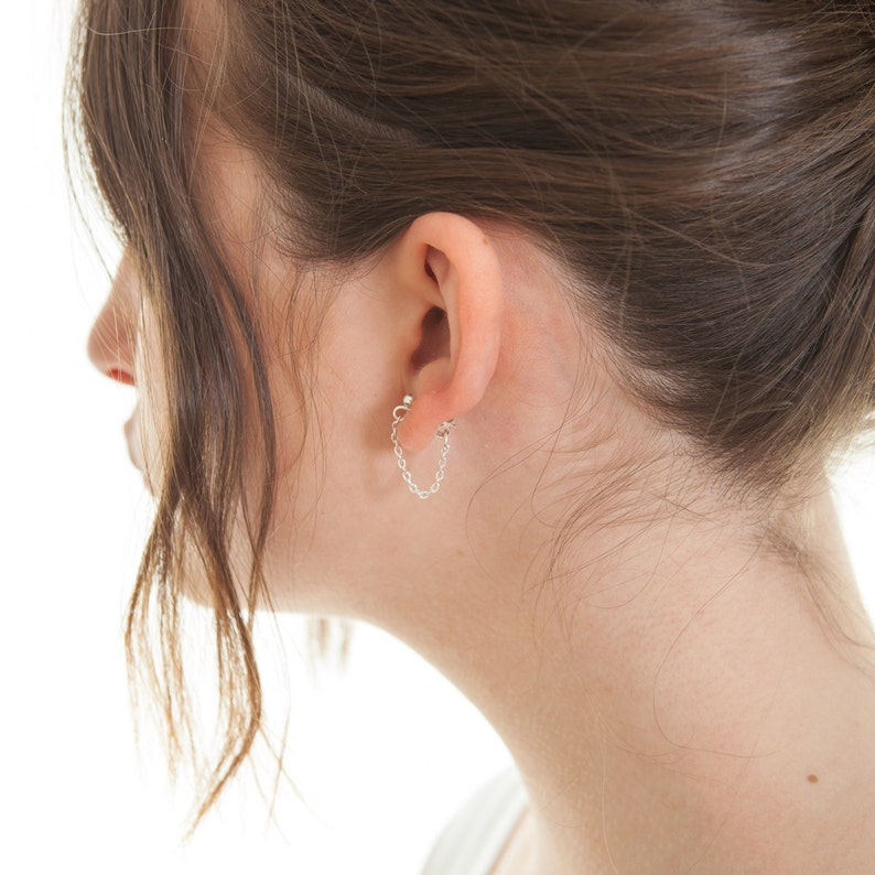 May include: A close-up of a person's ear with a silver chain earring. The chain is attached to a small stud in the earlobe.