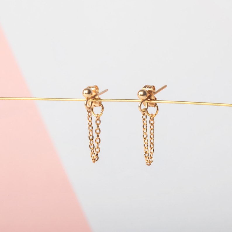 May include: A pair of gold-toned earrings with a small ball at the top and a delicate chain dangling down.