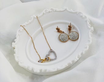 Puede incluir: Un collar dorado con un colgante en forma de corazón y un símbolo de infinito, junto con pendientes redondos a juego, exhibidos en un plato blanco adornado. La joyería está adornada con piedras claras y brillantes, sobre un fondo de tela blanca.