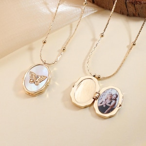 Puede incluir: Dos collares dorados con colgantes ovalados. Uno presenta un diseño de mariposa, el otro se abre para revelar una pequeña fotografía. Los collares se muestran sobre una superficie de color claro.