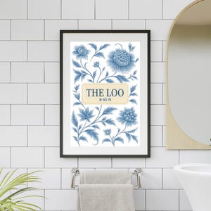 May include: Framed wall art featuring a blue floral design with the text "THE LOO" in a cream-colored rectangle. The artwork is in a black frame and hangs above a towel rack in a bathroom setting.