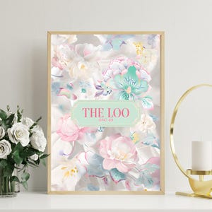 Op de afbeelding: Een ingelijste bloemenprint met een ontwerp in aquarel-stijl. Het kunstwerk toont pastelroze, witte en turquoise bloemen tegen een lichte achtergrond. De tekst "THE LOO @NO 49" staat op een mintgroen label.