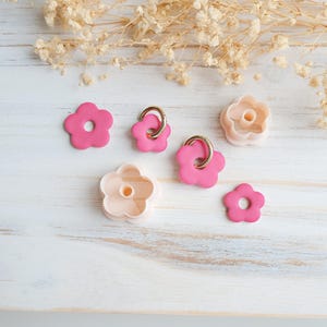 Op de afbeelding: Een verzameling roze bloemvormige items, waaronder oorbellen met gouden ringen en koekjessnijders, gerangschikt op een wit houten oppervlak. Gedroogde bloemaccenten op de achtergrond cre&euml;ren een zachte, pastelkleurige esthetiek.