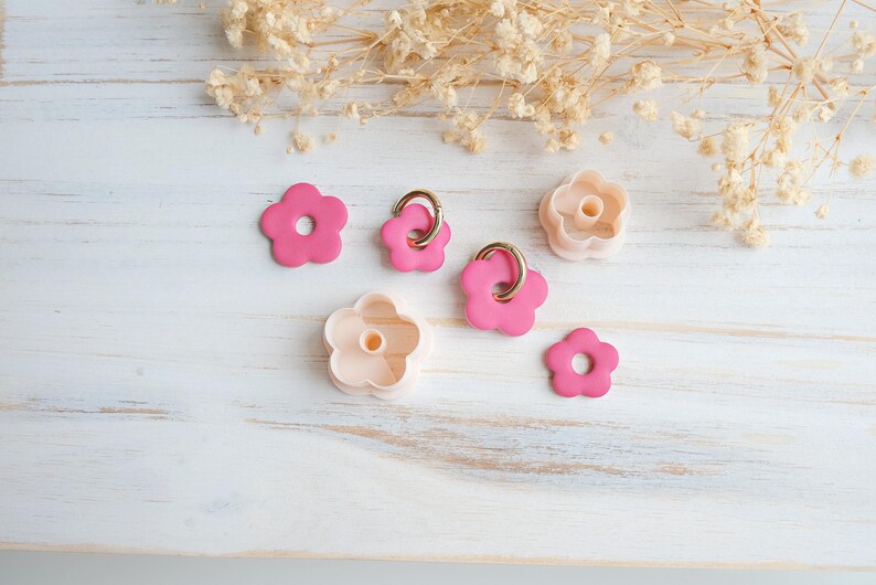 Op de afbeelding: Een verzameling roze bloemvormige items, waaronder oorbellen met gouden ringen en koekjessnijders, gerangschikt op een wit houten oppervlak. Gedroogde bloemaccenten op de achtergrond cre&euml;ren een zachte, pastelkleurige esthetiek.
