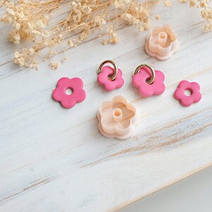 Op de afbeelding: Een verzameling roze bloemvormige polymeerklei uitsteekvormen en oorbellen met goudkleurige ringen. De uitsteekvormen zijn in twee roze tinten en de oorbellen zijn helderder roze. De items zijn gerangschikt op een wit houten oppervlak met gedroogde bloemen.