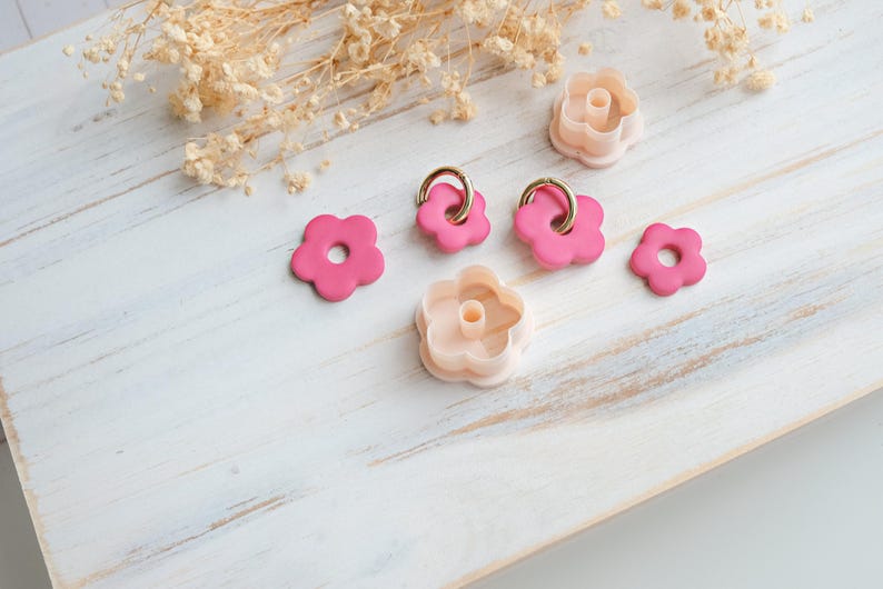 Op de afbeelding: Een verzameling roze bloemvormige polymeerklei uitsteekvormen en oorbellen met goudkleurige ringen. De uitsteekvormen zijn in twee roze tinten en de oorbellen zijn helderder roze. De items zijn gerangschikt op een wit houten oppervlak met gedroogde bloemen.