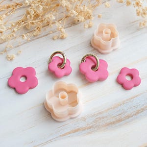 Op de afbeelding: Roze bloemvormige oorbellen met gouden ringen en perzikkleurige koekjessnijders. De items zijn gerangschikt op een wit houten oppervlak, met gedroogde bloemenaccenten. Bloemen thema.