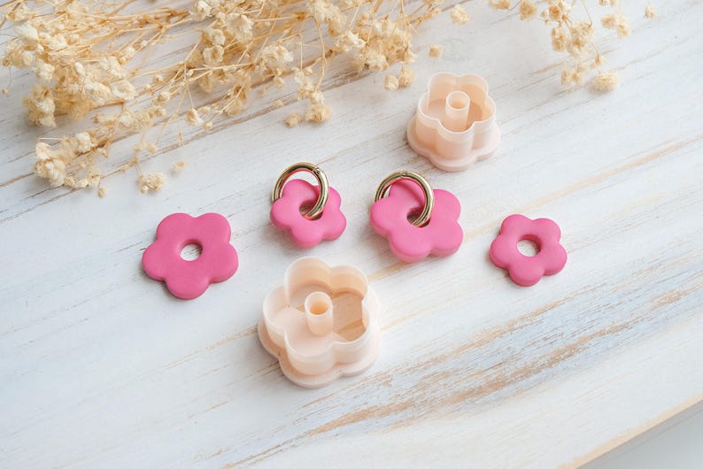 Op de afbeelding: Roze bloemvormige oorbellen met gouden ringen en perzikkleurige koekjessnijders. De items zijn gerangschikt op een wit houten oppervlak, met gedroogde bloemenaccenten. Bloemen thema.