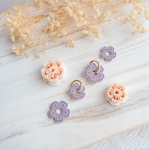 May include: A collection of floral-shaped craft supplies in shades of peach and lavender. Some flowers have gold-colored rings. The items are arranged on a white wooden surface, with dried flowers in the background.