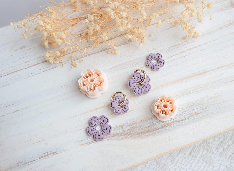 May include: A collection of floral-shaped craft supplies in shades of peach and lavender. Some flowers have gold-colored rings. The items are arranged on a white wooden surface, with dried flowers in the background.
